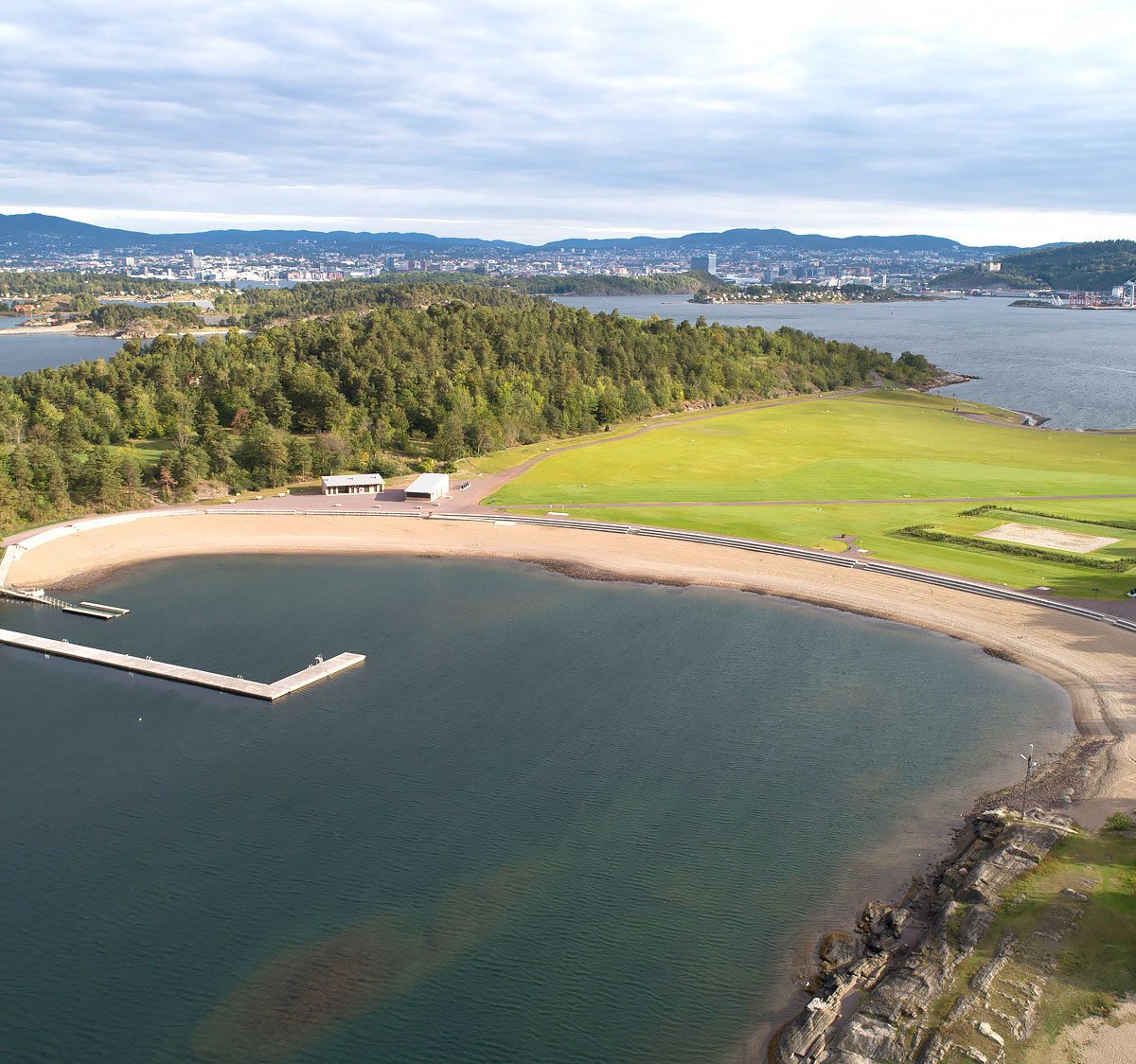 Dronebilde av Nye Langøyene viser en u-formet tilrettelagt badestrand i forkant av en gjennomgående gresslette, omgitt av løvskog. Utformet av landskapsarkitekter og et tverrfaglig teamFoto: Asplan Viak/Jostein Thorvaldsen