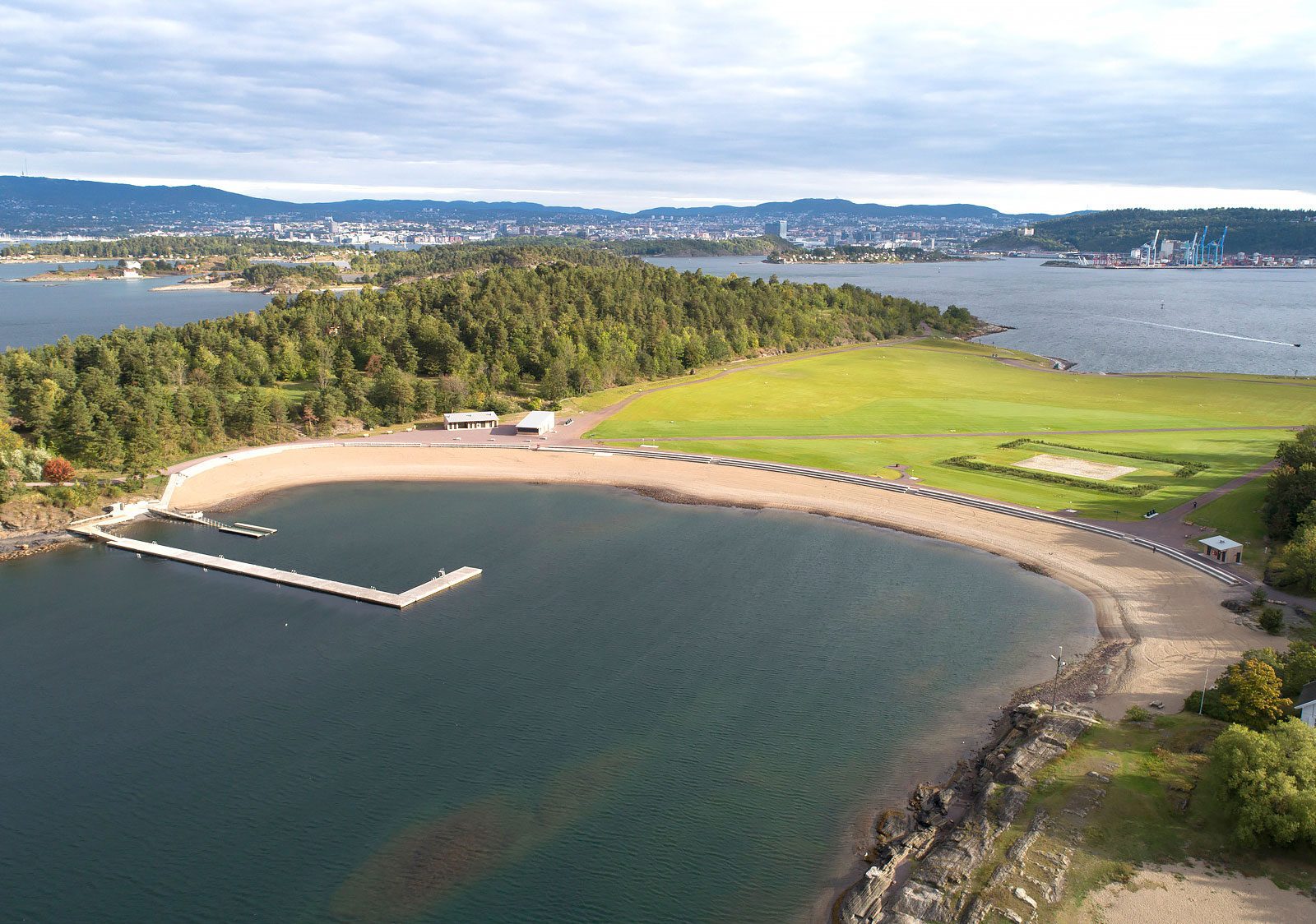 Dronebilde av Nye Langøyene viser en u-formet tilrettelagt badestrand i forkant av en gjennomgående gresslette, omgitt av løvskog. Utformet av landskapsarkitekter og et tverrfaglig teamFoto: Asplan Viak/Jostein Thorvaldsen