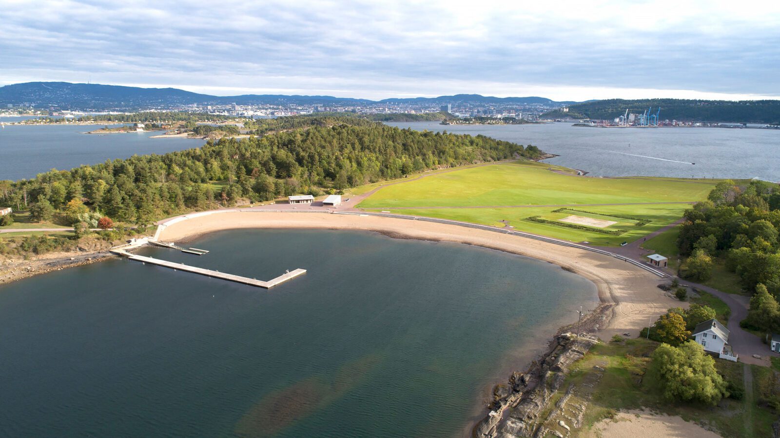 Dronebilde av Nye Langøyene viser en u-formet tilrettelagt badestrand i forkant av en gjennomgående gresslette, omgitt av løvskog. Foto: Asplan Viak/Jostein Thorvaldsen