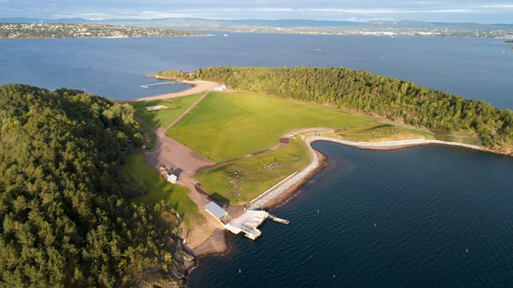 Dronebilde av Nye Langøyene viser en badestrand og en gjennomgående gresslette, omgitt av løvskog på begge sider. Foto: Asplan Viak/Jostein Thorvaldsen