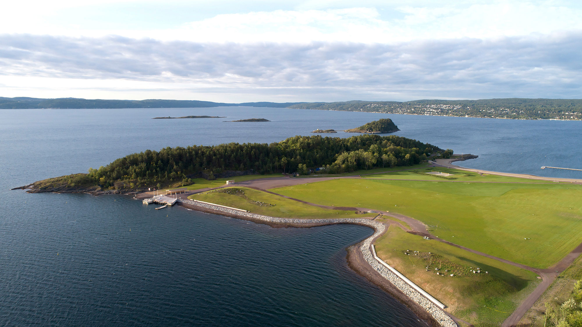 Dronebilde av Nye Langøyene viser strandpromenaden foran en gjennomgående gresslette, med løvskog i bakgrunnen. Foto: Asplan Viak/Jostein Thorvaldsen